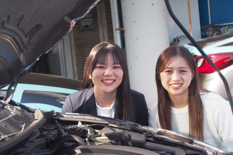 バンパーを開けた教習車越しにカメラに笑顔を向けている講師と在校生